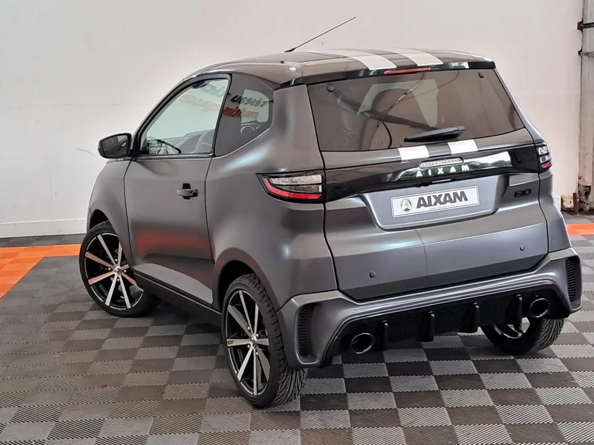 Vue 3/4 arrière droite d'une Aixam Ambition gris clair avec jantes alliage noires diamantées et bandes blanches sur le toit.