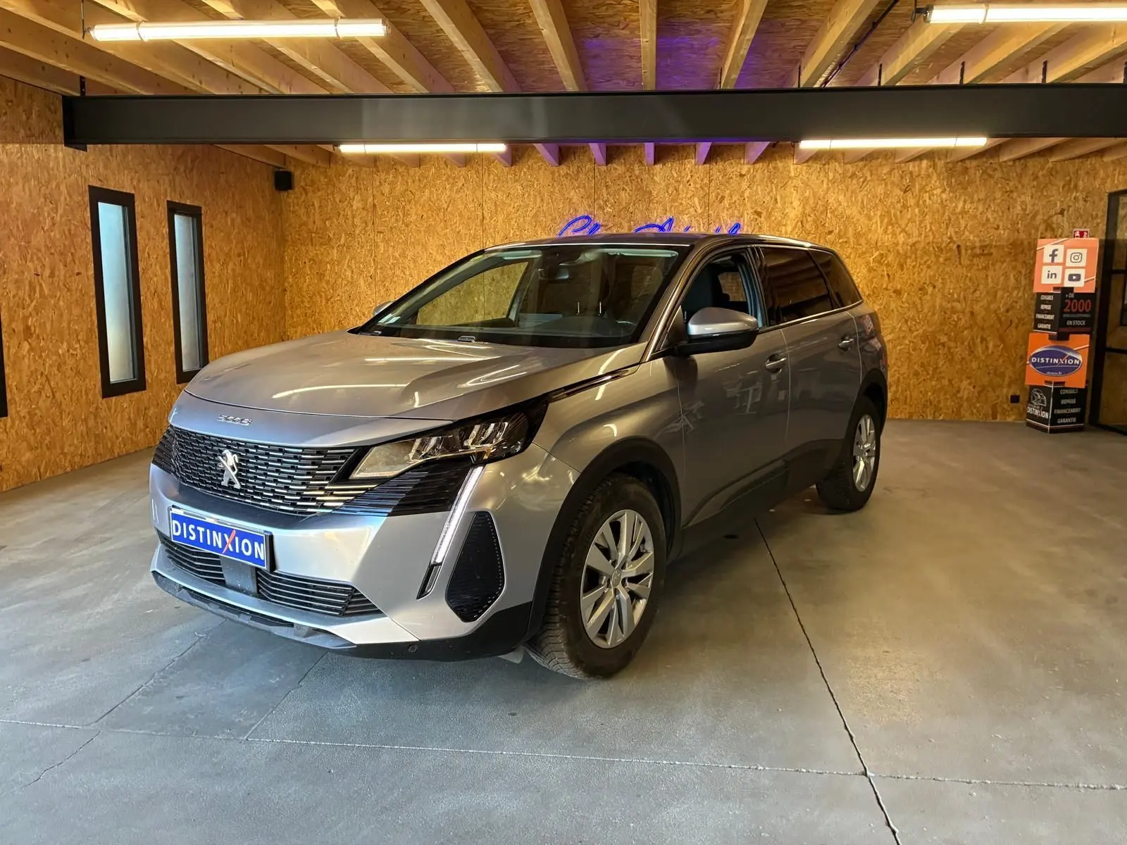 Peugeot 5008 gris en 3/4 avant droit dans un showroom avec éclairage intérieur et mur en bois.