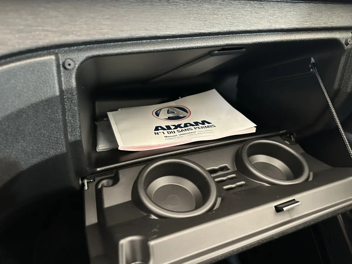 Gros plan sur la boîte à gants ouverte de l'AIXAM AMBITION E CITY PACK avec manuel utilisateur visible à l'intérieur.
