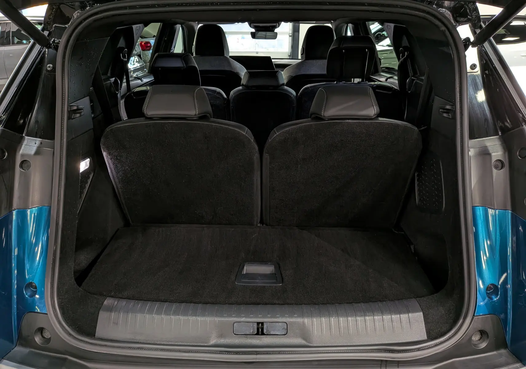 Coffre ouvert vu de l’arrière d’un Peugeot 5008 Hybrid bleu Obsession avec intérieur noir et sièges rabattus partiellement.