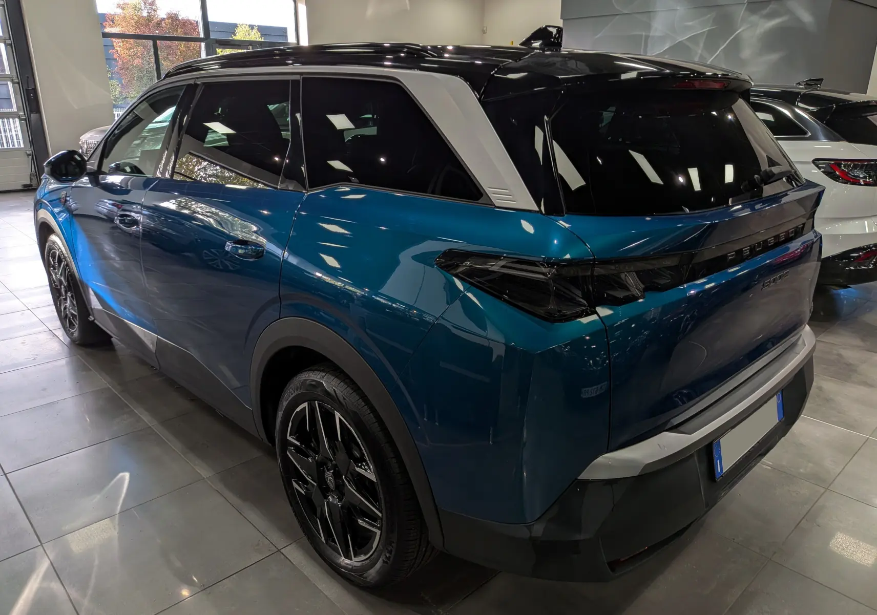 Vue 3/4 arrière droite du Peugeot 5008 Hybrid 145 GT bleu Obsession avec toit noir et jantes noires en intérieur showroom.