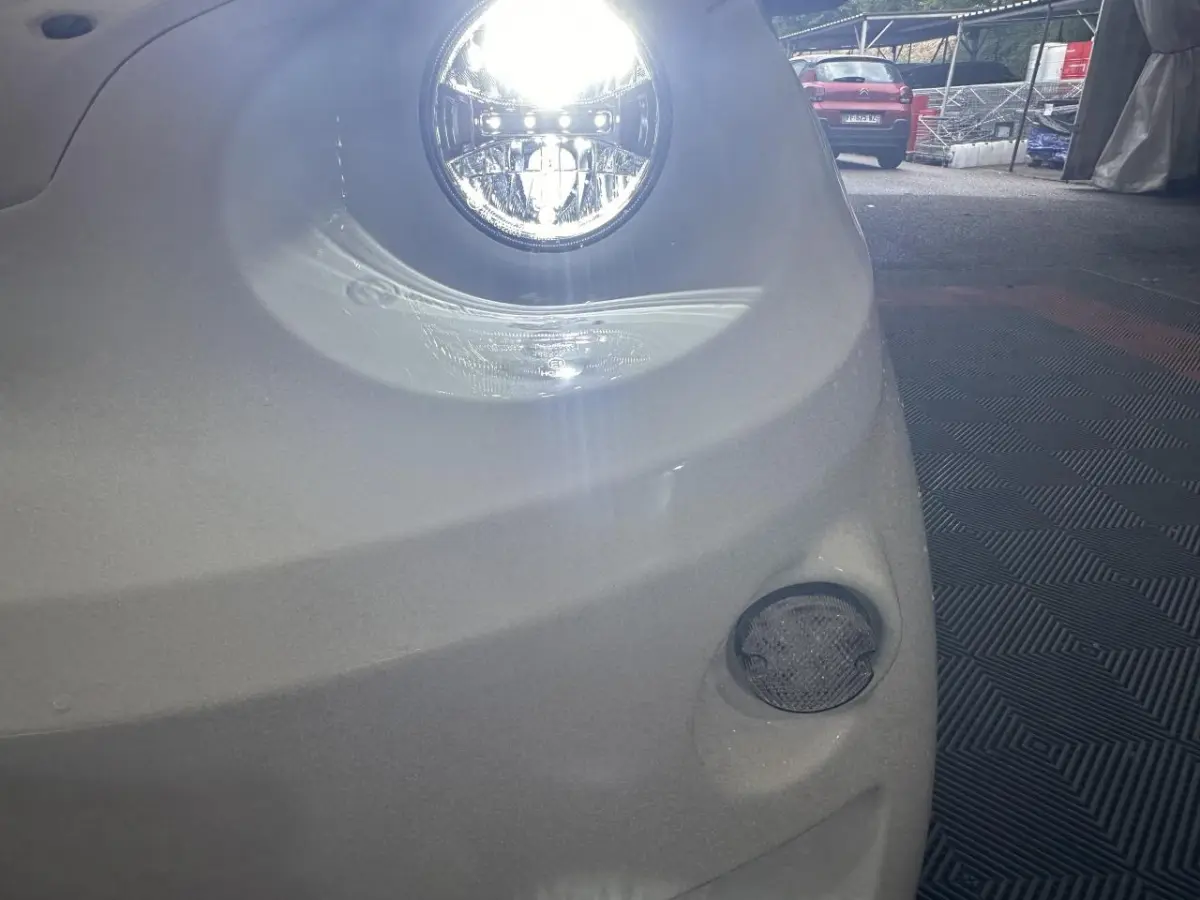 Gros plan sur le phare avant droit allumé d'une Aixam Minauto E Access blanche, avec un feu antibrouillard visible en bas.