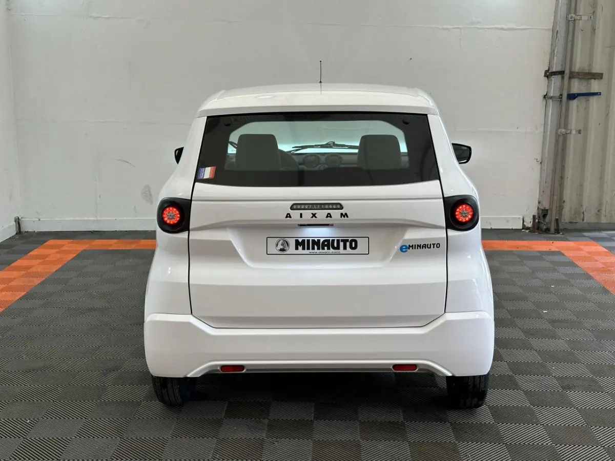 Vue arrière d'une AIXAM MINAUTO E ACCESS blanche 2026 avec feux ronds et logo MINAUTO visible sur un sol à damier noir et orange.