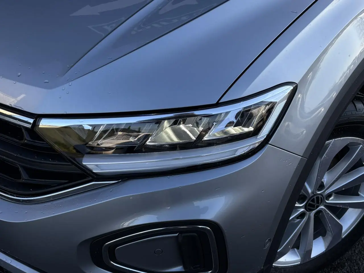 Gros plan sur l'avant droit du Volkswagen T-Roc gris Indium, mettant en valeur le phare LED et la roue alliage.