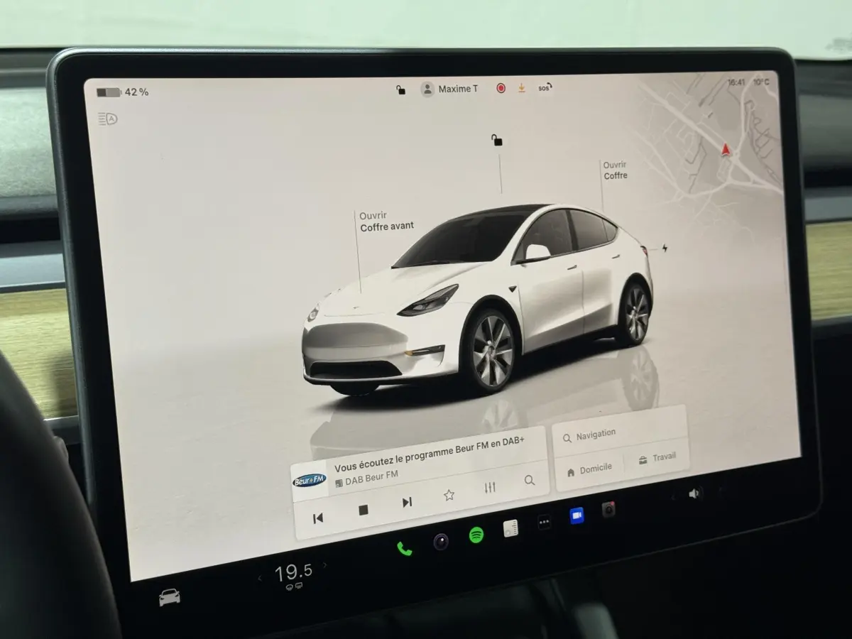 Vue 3/4 avant d’un Tesla Model Y blanc affiché sur l’écran tactile central avec options d’ouverture des coffres.
