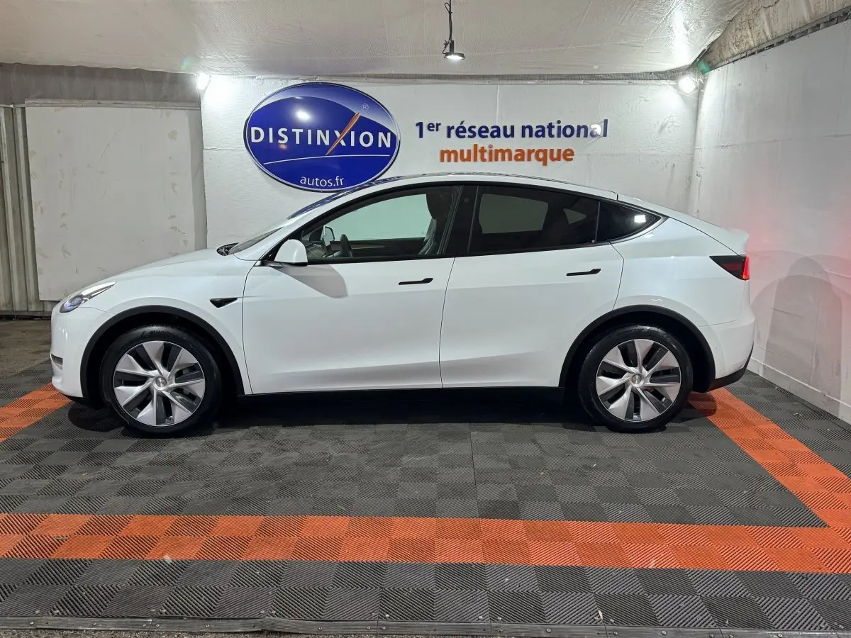 Vue de profil côté gauche d'une Tesla Model Y blanc nacré, stationnée en intérieur sur sol noir et orange.
