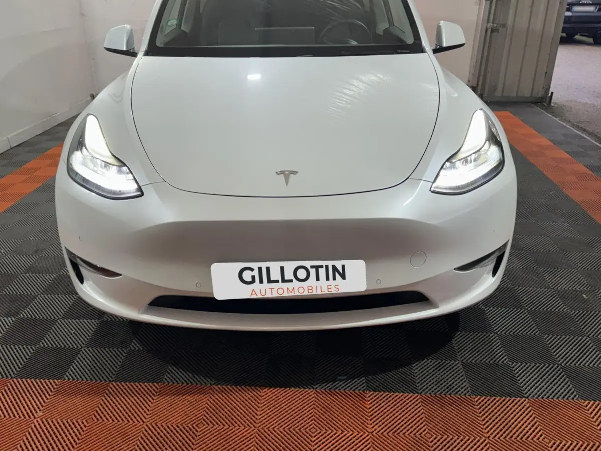 Vue avant d'une Tesla Model Y blanche avec phares LED allumés, sur sol damier noir et orange en intérieur.