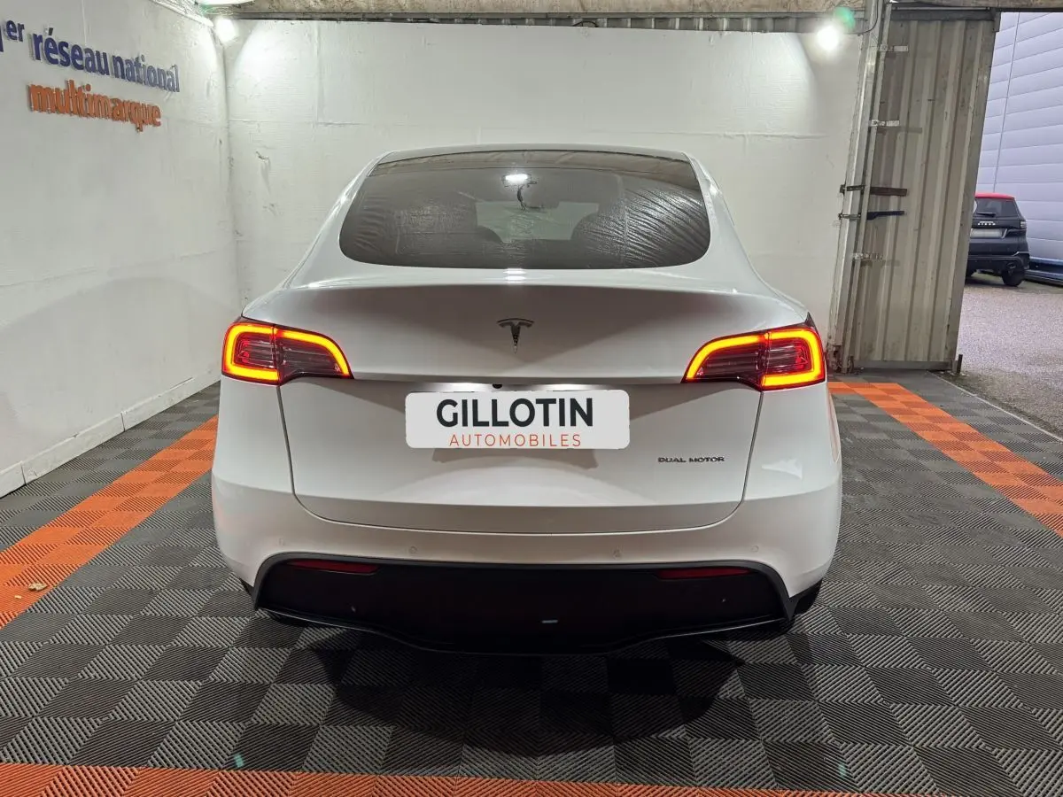 Vue arrière d'une Tesla Model Y blanc nacré avec feux arrière allumés dans un garage éclairé.