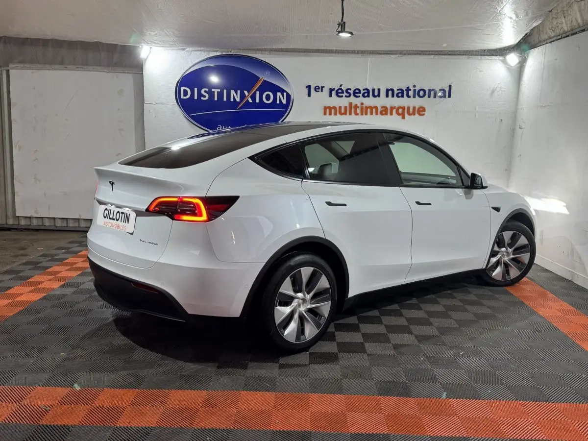 Vue 3/4 arrière droite d'une Tesla Model Y blanc nacré avec feux arrière allumés dans un showroom.