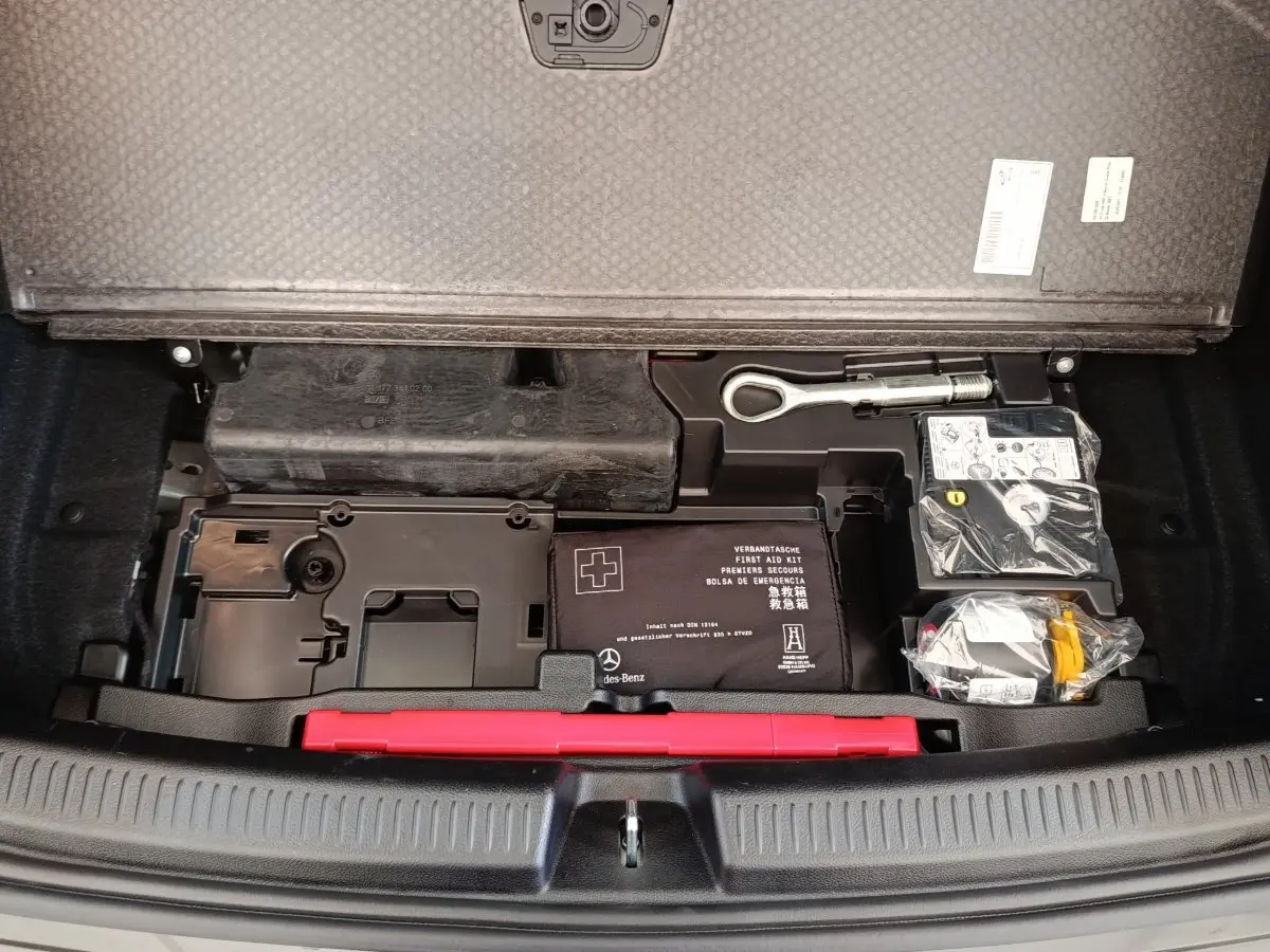 Vue plongeante sur le coffre ouvert d'une Mercedes Classe A noire, montrant la trousse de secours et outils rangés sous la tablette.