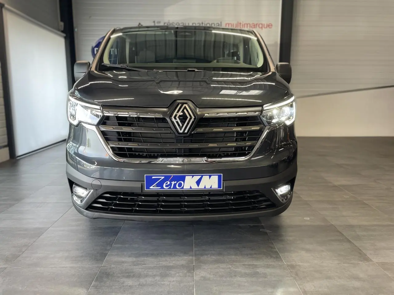 Vue frontale du Renault Trafic Fourgon gris Comète 2025 avec phares Full LED allumés en intérieur showroom.