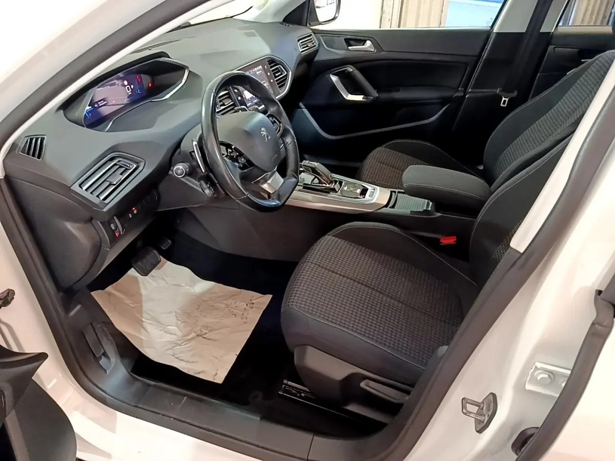 Intérieur avant droit de la Peugeot 308 blanche, montrant le volant cuir, console centrale et sièges noirs tissu.