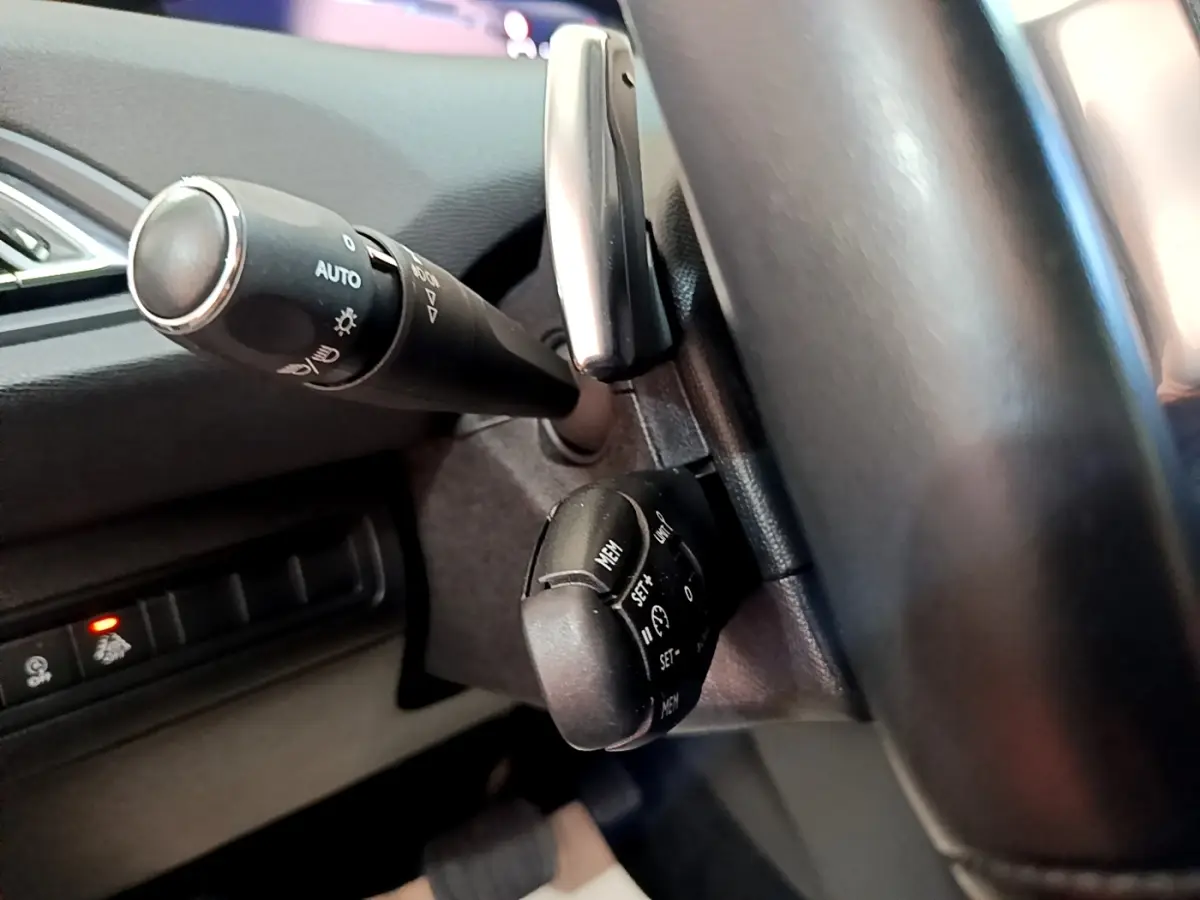 Gros plan sur les commandes au volant noir de la Peugeot 308 blanche, mettant en valeur les fonctions de régulateur et éclairage.