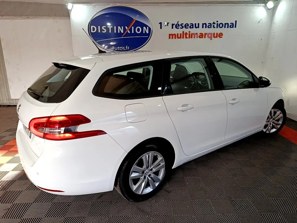 Peugeot 308 break blanc vue 3/4 arrière droit, feux arrière allumés, stationné en intérieur sur sol à motifs.