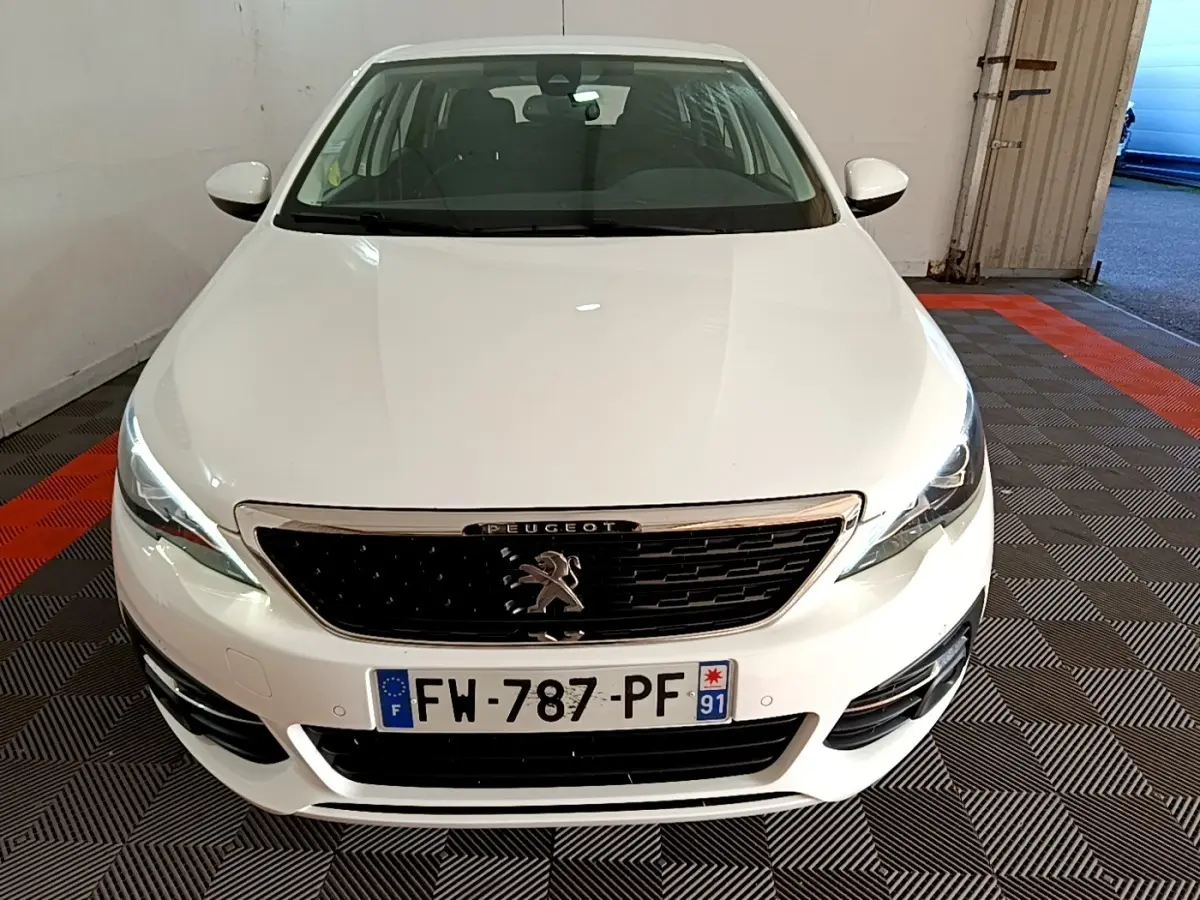 Vue de face d'une Peugeot 308 blanche avec calandre noire et phares LED allumés dans un garage.