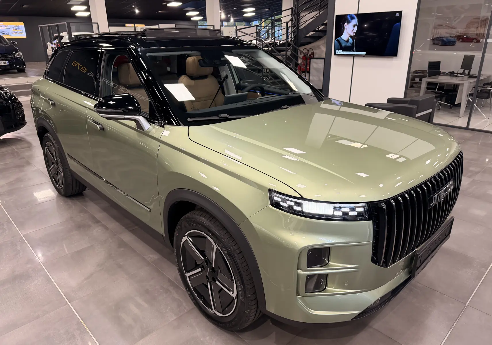 Vue 3/4 avant d’un SUV JAECOO J7 hybride rechargeable gris olive avec toit noir et intérieur cuir brun en showroom.
