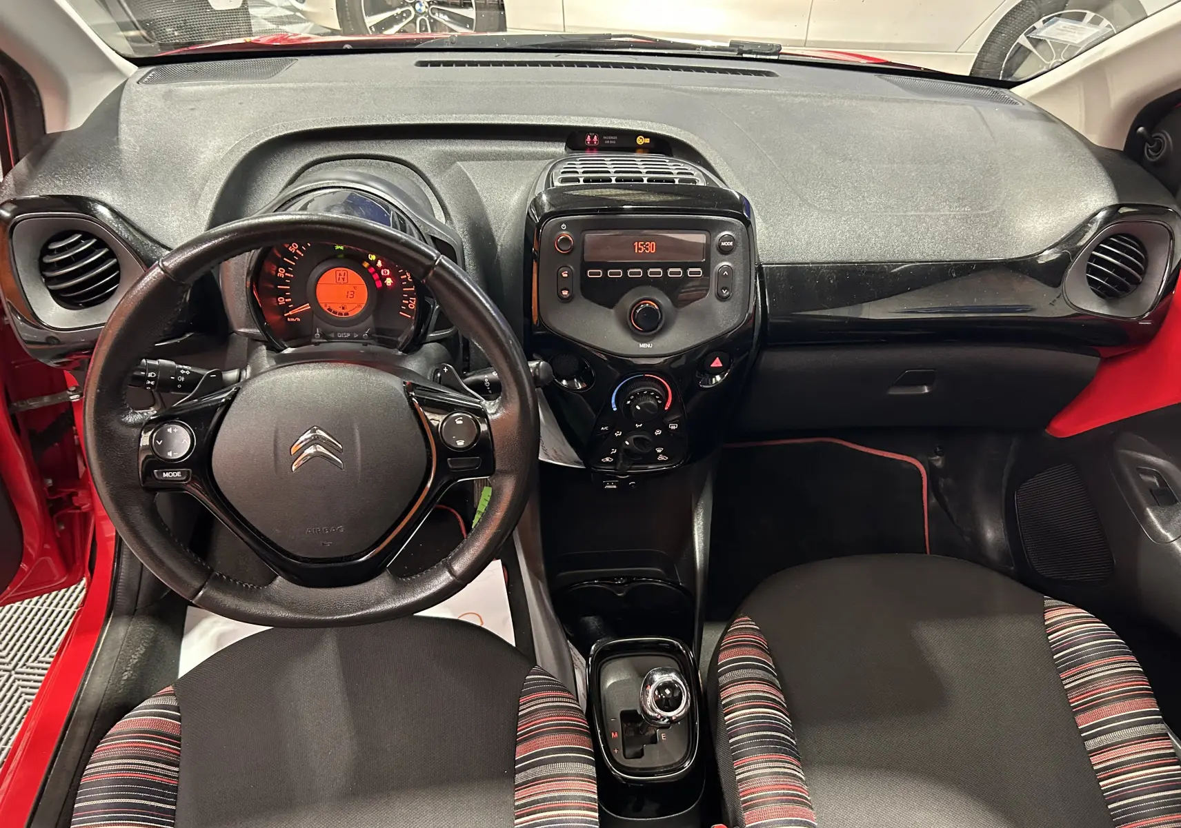 Vue intérieure avant du tableau de bord et volant noir d’une Citroën C1 rouge avec sièges tissu à rayures colorées.