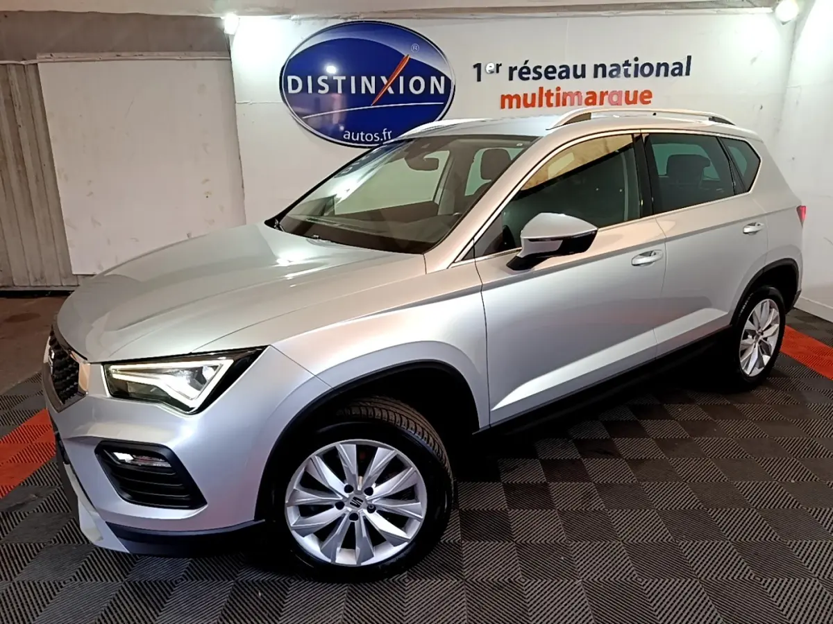 Vue 3/4 avant droit d’un SEAT Ateca gris clair 2022, avec jantes alliage et barres de toit chromées, en intérieur showroom.