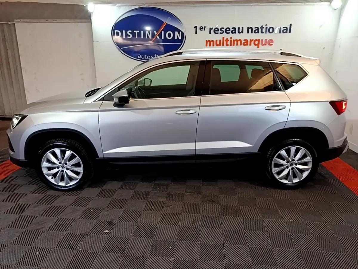 Vue de profil côté gauche d’un SEAT Ateca gris clair, avec jantes alliage et barres de toit chromées, en intérieur showroom.