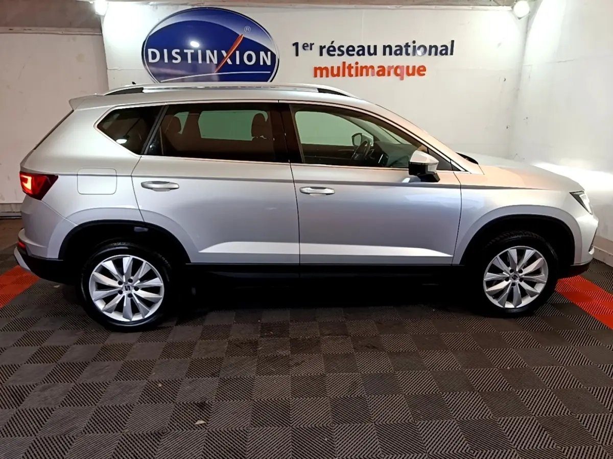 Vue latérale droite d'un SEAT Ateca gris clair 2022 dans un showroom avec logo Distinxion en arrière-plan.