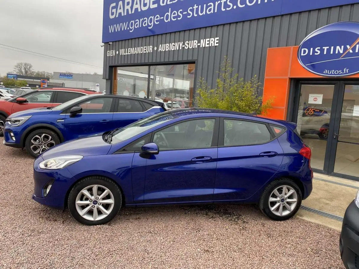 Vue de profil côté gauche d'une Ford Fiesta 1.1i Trend bleu foncé garée devant un garage, sur sol gravillonné.