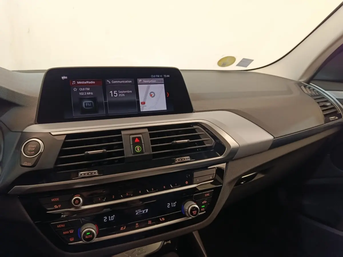 Tableau de bord intérieur de BMW X3 gris clair, vue de face mettant en valeur l’écran tactile et les commandes climatisation.
