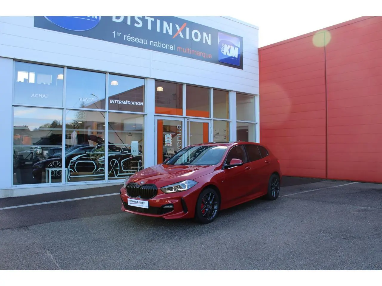 BMW Série 1 118d rouge Melbourne métallisé vue 3/4 avant droit, avec jantes alliage et becquet arrière M Sport.