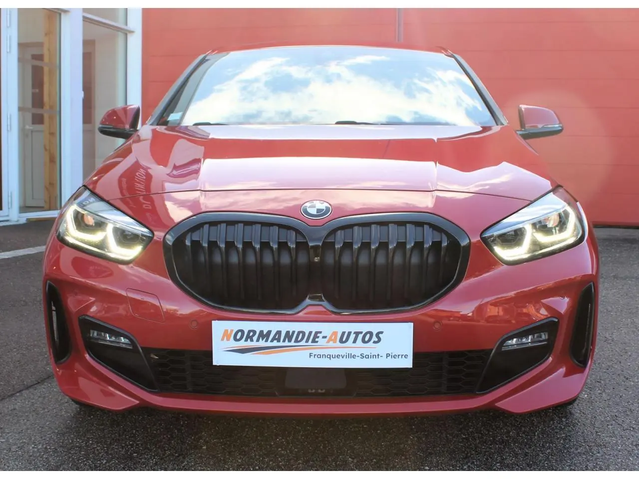 Vue avant d'une BMW Série 1 118d rouge Melbourne avec calandre noire et feux LED allumés.