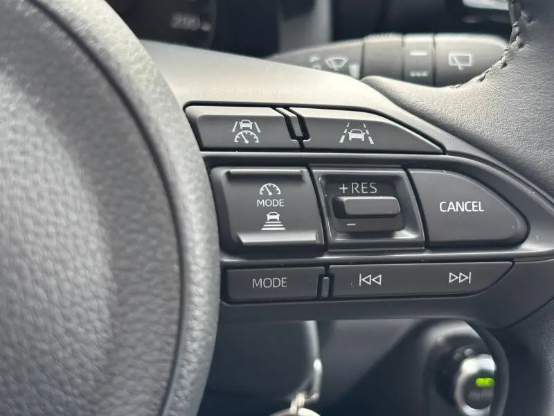 Gros plan sur les commandes au volant noir de la Toyota Yaris 116h Dynamic 5p MC24 blanche, avec boutons de régulateur et aide à la conduite.