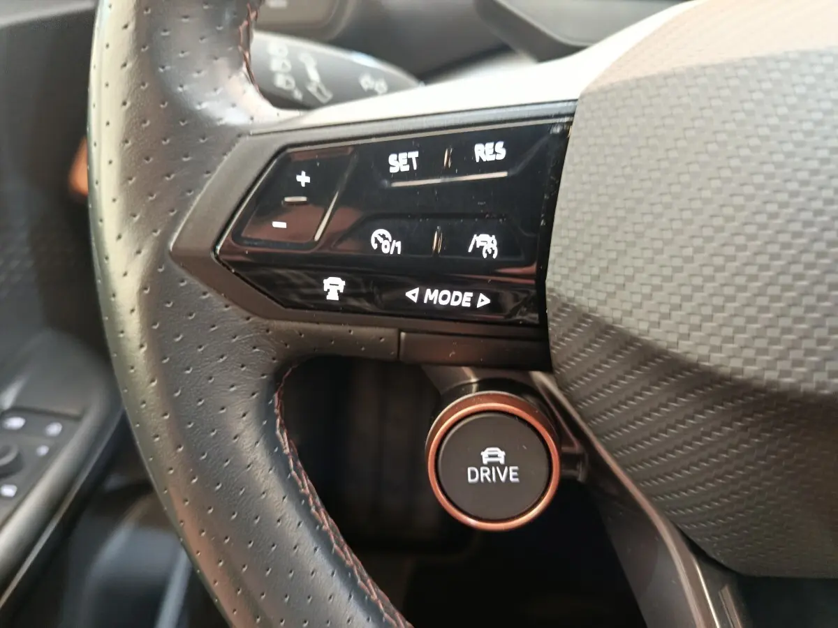 Gros plan sur les commandes du volant cuir noir perforé du Cupra Born 230ch, bouton Drive et réglages du régulateur.