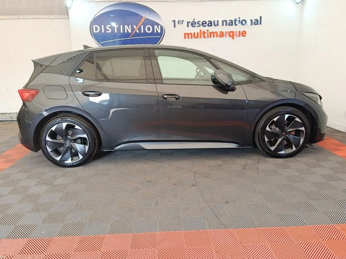 Vue de profil côté gauche d'une CUPRA BORN 230ch gris foncé avec jantes noires et toit spoiler en intérieur showroom.