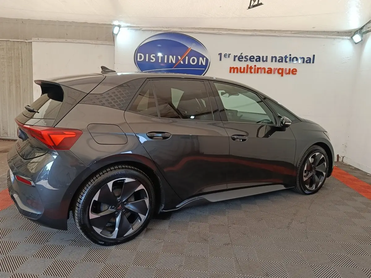 Vue latérale droite d'une CUPRA BORN 230ch gris foncé avec jantes noires et toit avec spoiler en intérieur showroom.