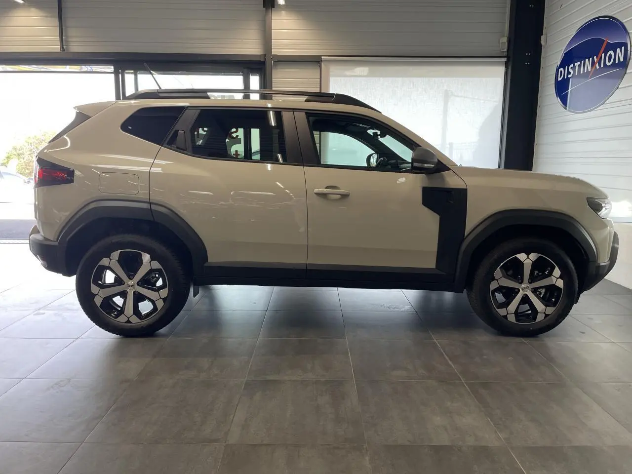 Profil droit du SUV Dacia Duster beige avec jantes alliage et barres de toit noires en intérieur showroom.