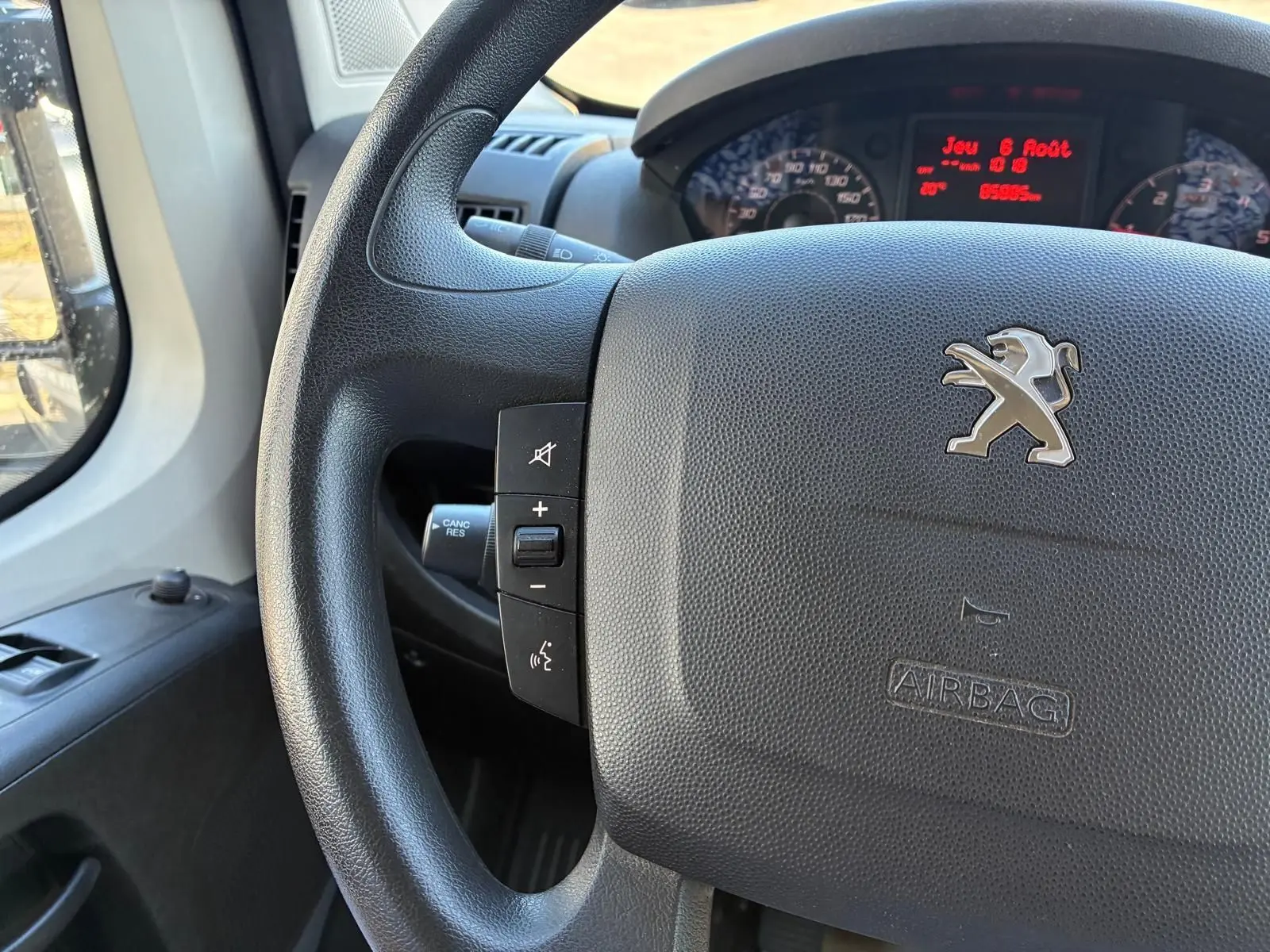 Gros plan sur le volant noir du Peugeot Boxer 2019 avec commandes intégrées et tableau de bord digital rouge en arrière-plan.