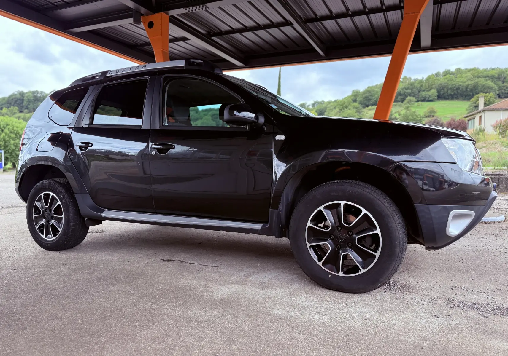 Vue de profil côté gauche d'un Dacia Duster noir métal 2017 sous abri avec jantes bi-ton et barres de toit.
