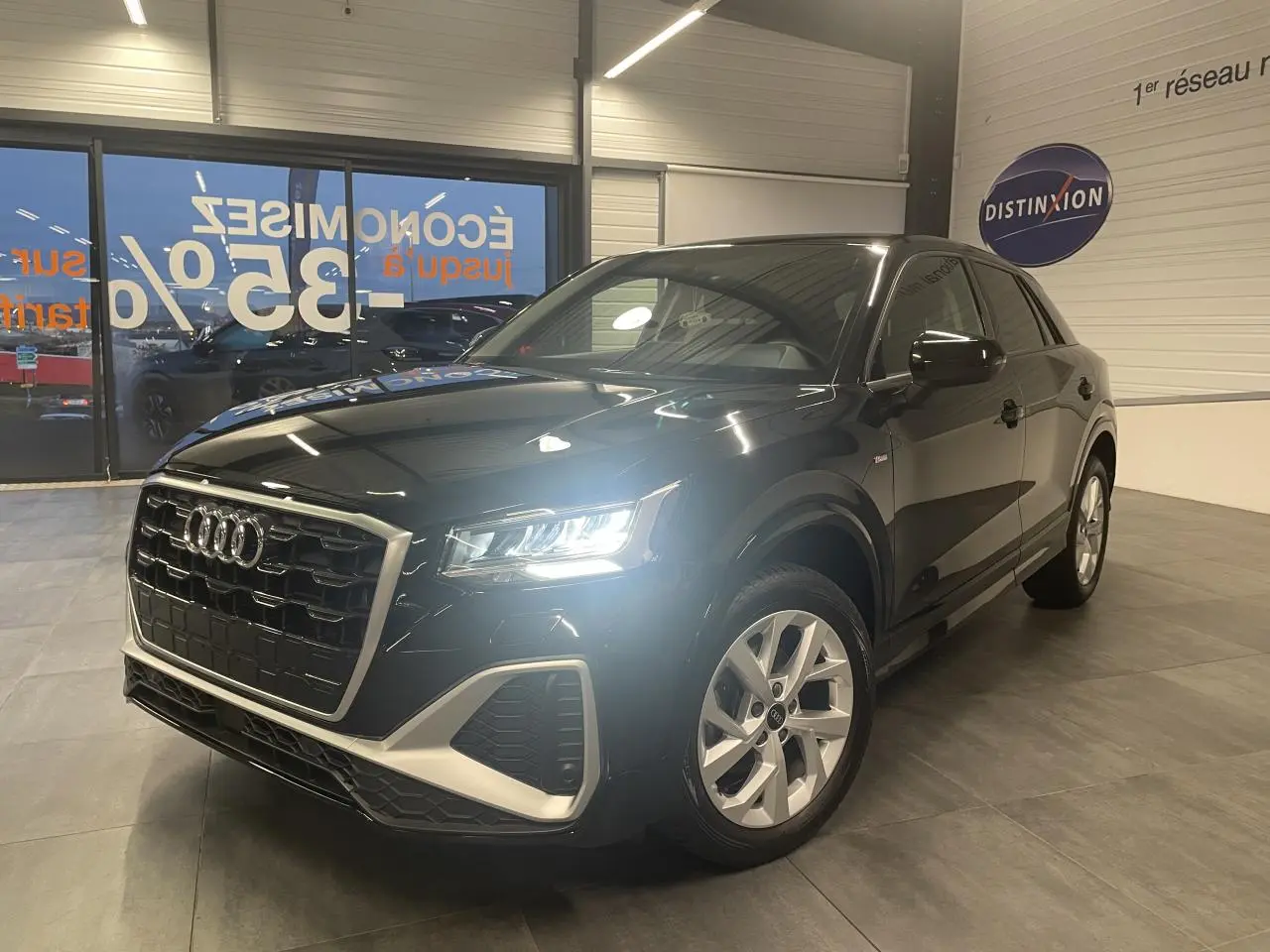 Audi Q2 noir métallisé vue 3/4 avant droit en intérieur, avec phares LED allumés et jantes alliage visibles.