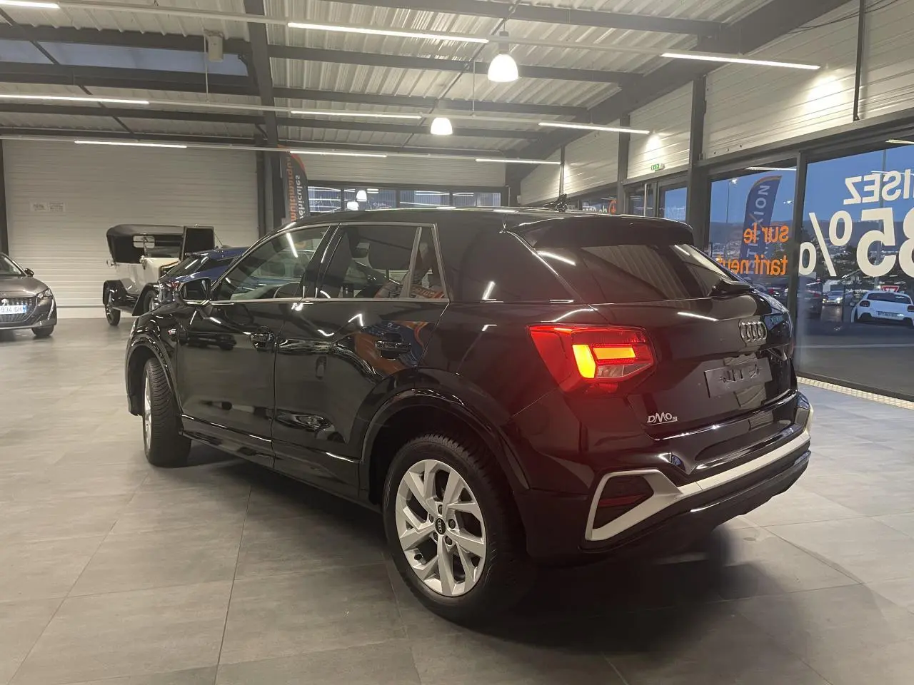 Audi Q2 noir métallisé vue 3/4 arrière droit en intérieur avec feux arrière LED allumés et jantes alliage.