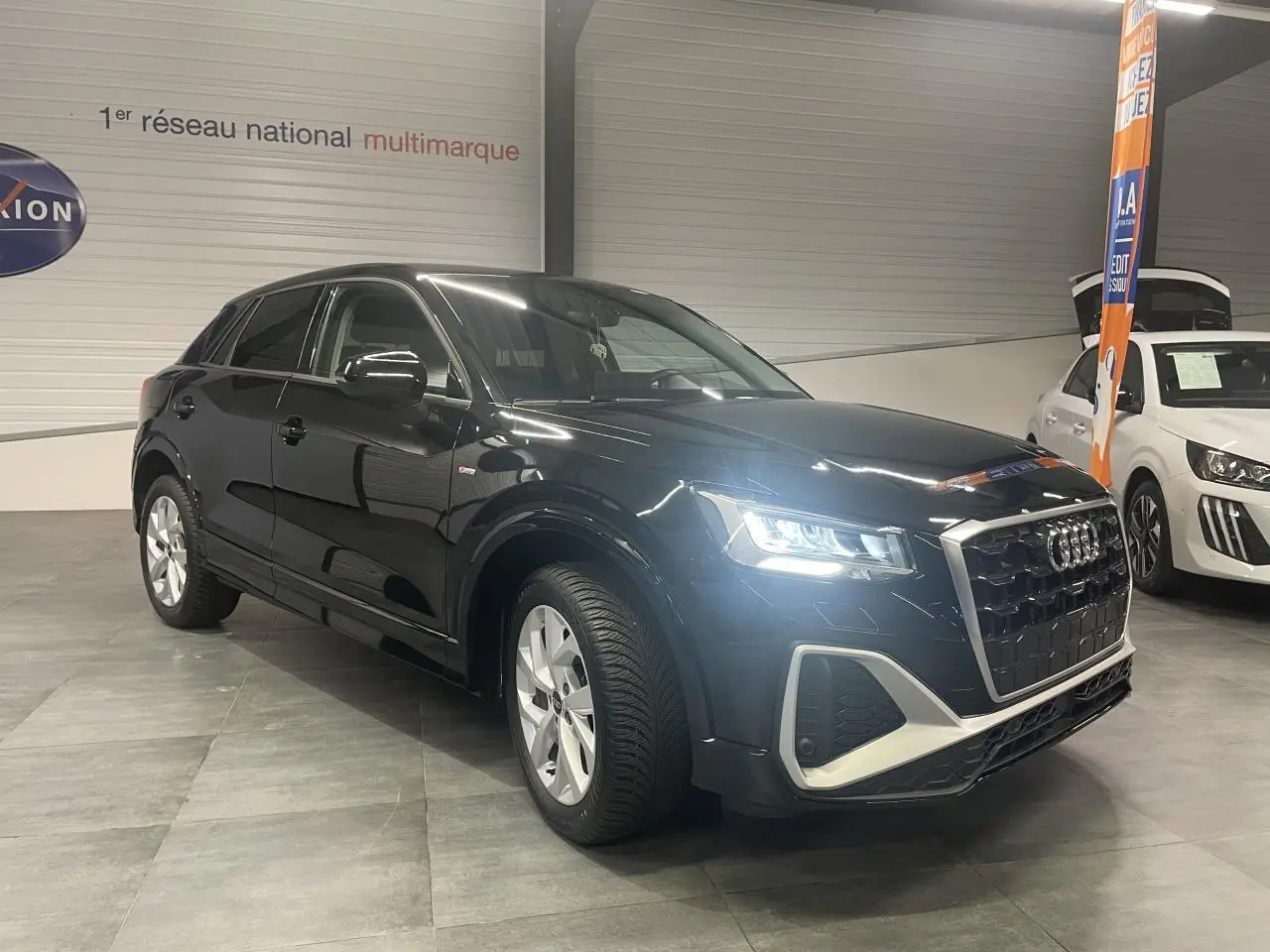 Audi Q2 noir métallisé vue 3/4 avant droit avec phares allumés dans un showroom intérieur.