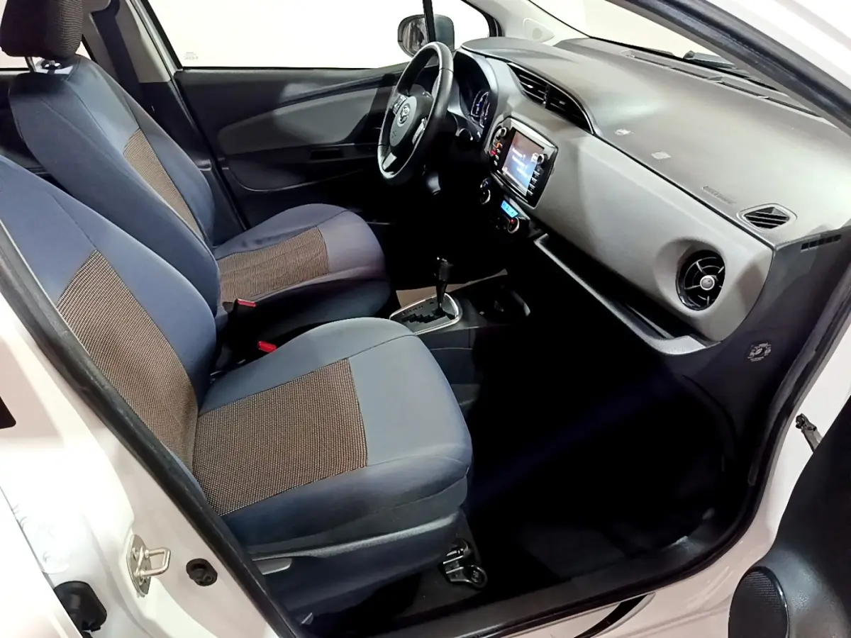 Vue intérieure côté conducteur de la Toyota Yaris blanche 2019, montrant sièges tissu bicolore et tableau de bord avec écran tactile.