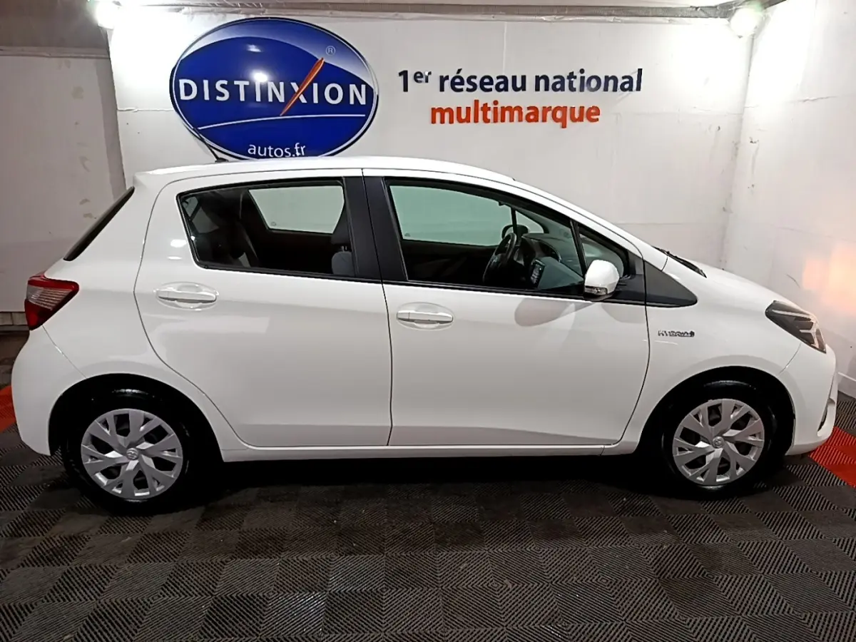 Vue de profil côté gauche d'une Toyota Yaris blanche hybride 2019, avec jantes et rétroviseurs blancs dans un showroom.