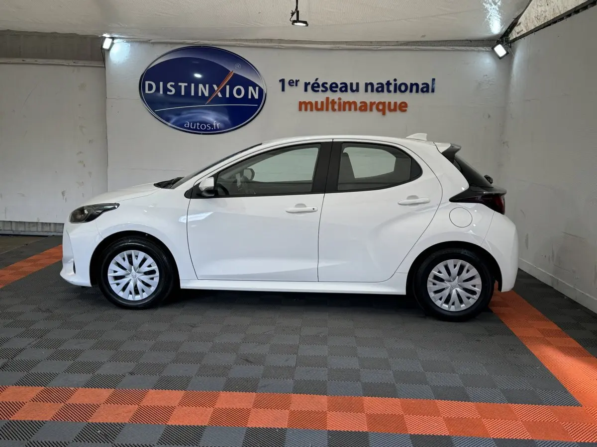 Profil côté gauche d'une Toyota Yaris hybride blanche 2022, avec jantes pleines et feux arrière noirs dans un showroom.