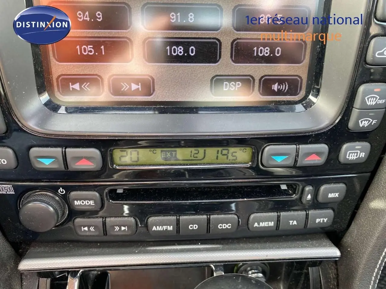 Gros plan sur la console centrale noire de la Jaguar XJ6 2.7 V6 Biturbo de 2006, montrant l'autoradio et la climatisation digitale.