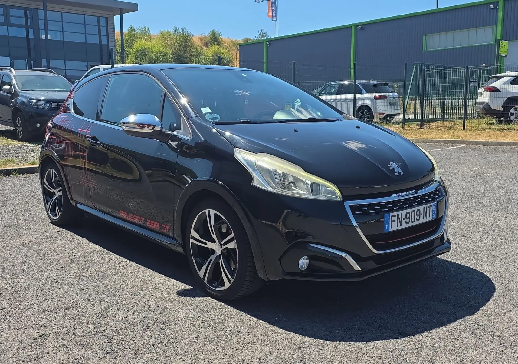Peugeot 208 GTI noire vue 3/4 avant droit avec jantes alliage 17” et stickers rouges sur la portière arrière.