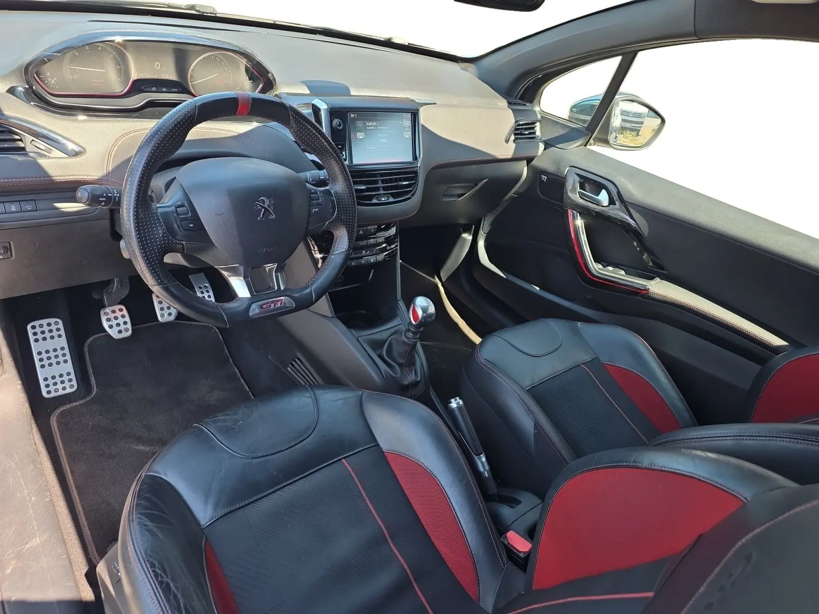 Vue intérieure côté conducteur de la Peugeot 208 GTI 2015 avec sellerie cuir noir et rouge, volant sport et levier de vitesse manuel.