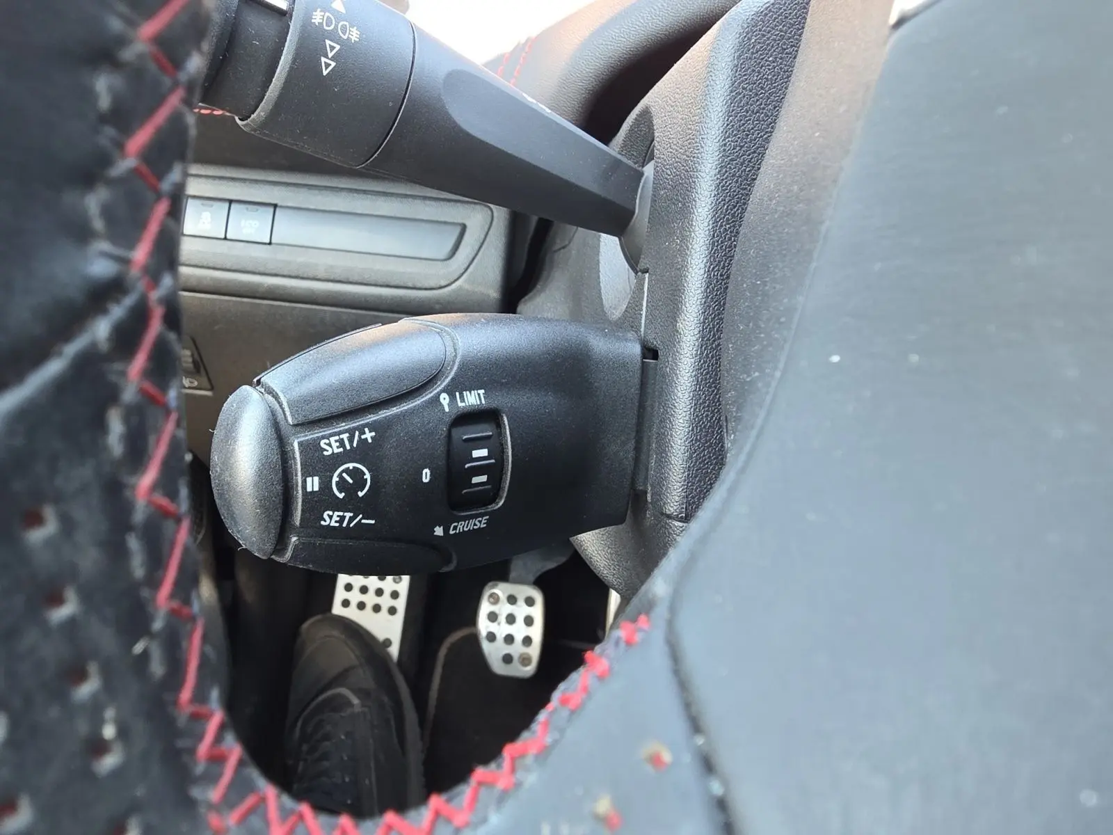 Gros plan sur la commande de régulateur de vitesse à gauche du volant cuir noir avec surpiqûres rouges dans une Peugeot 208 GTI noire.