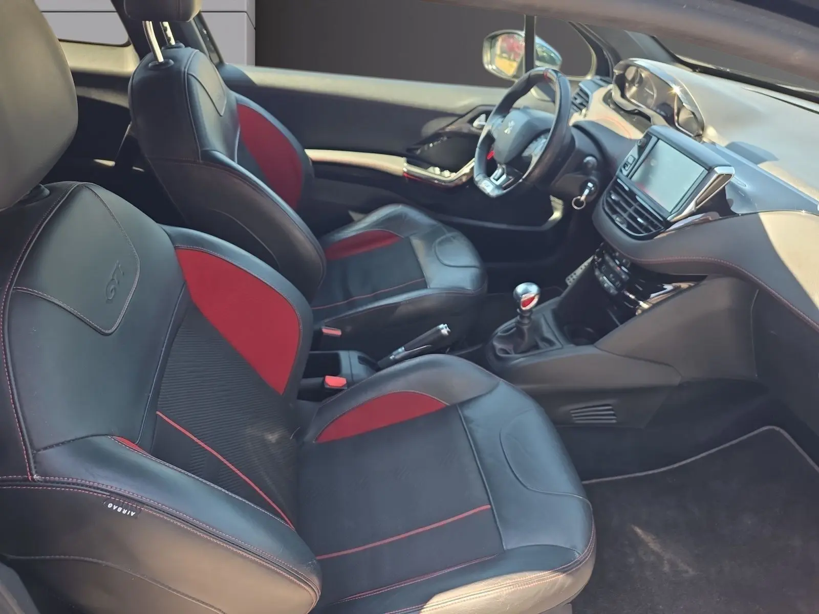 Vue intérieure côté conducteur de la Peugeot 208 GTI 2015, sièges baquets cuir noir et rouge, volant sport et tableau de bord moderne.