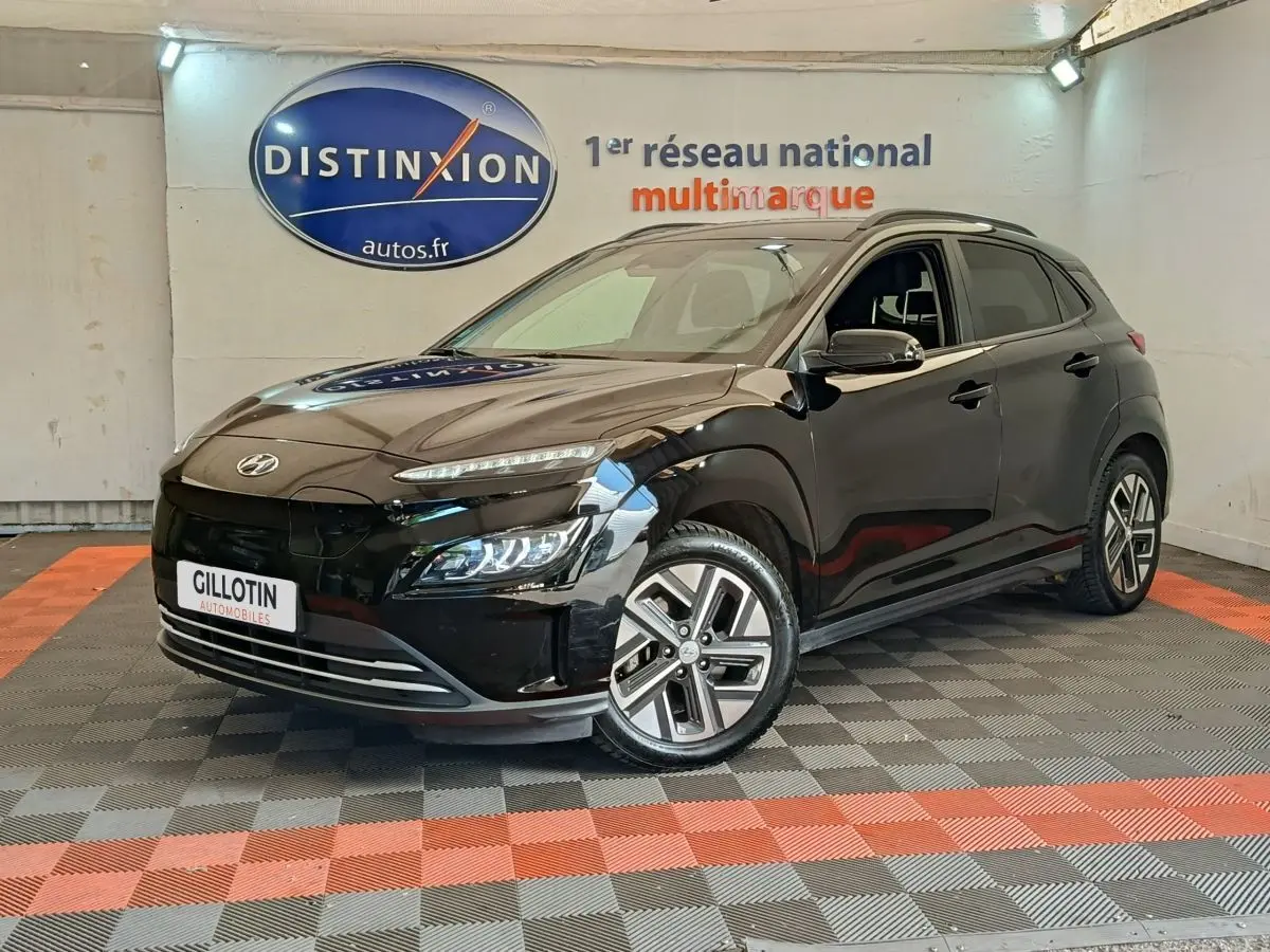 Hyundai Kona électrique noir en 3/4 avant droit, avec jantes spécifiques et feux LED allumés en intérieur showroom.