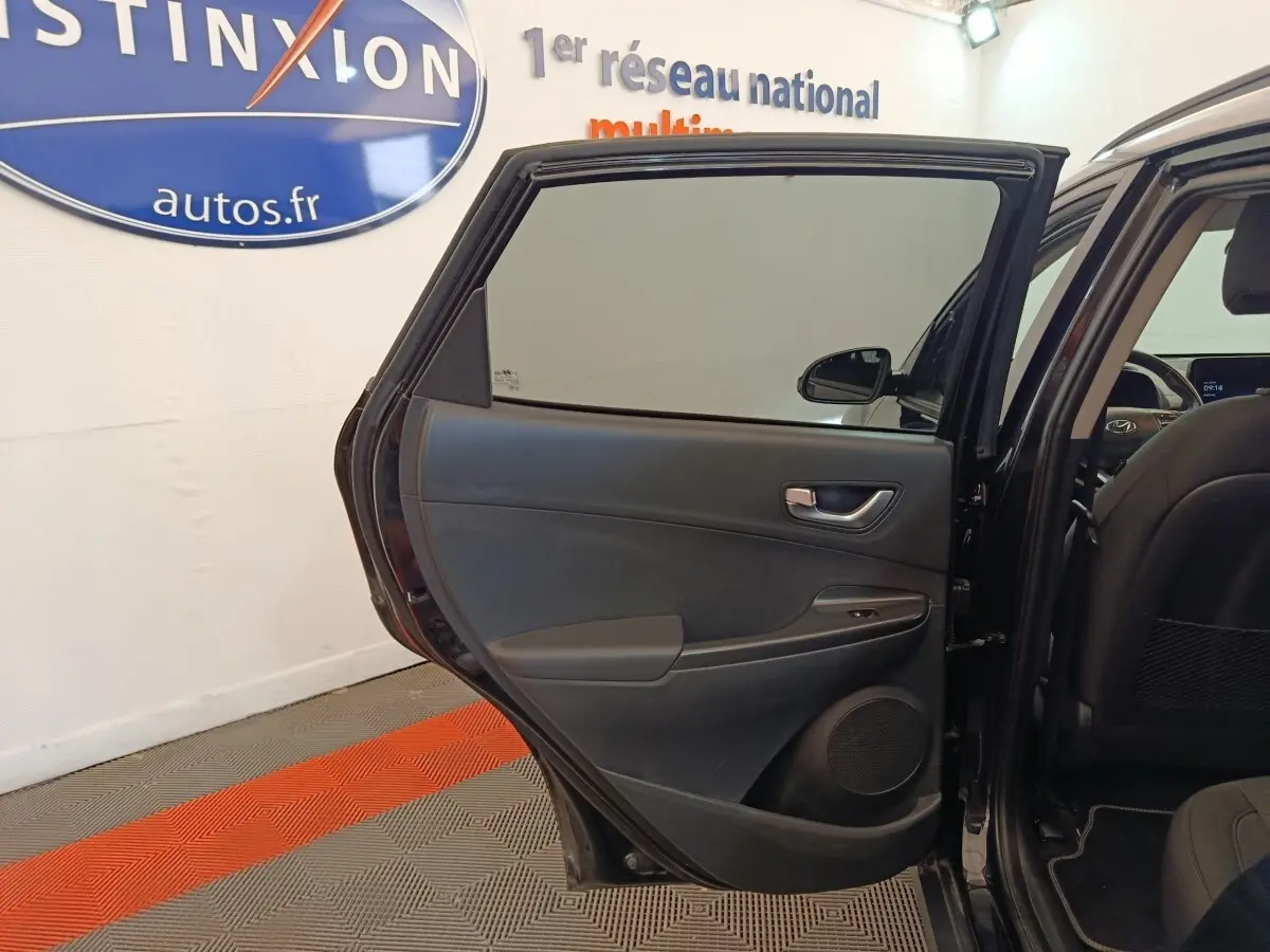 Vue intérieure de la porte arrière gauche ouverte d'une Hyundai Kona noire, avec siège arrière et tableau de bord partiellement visibles.