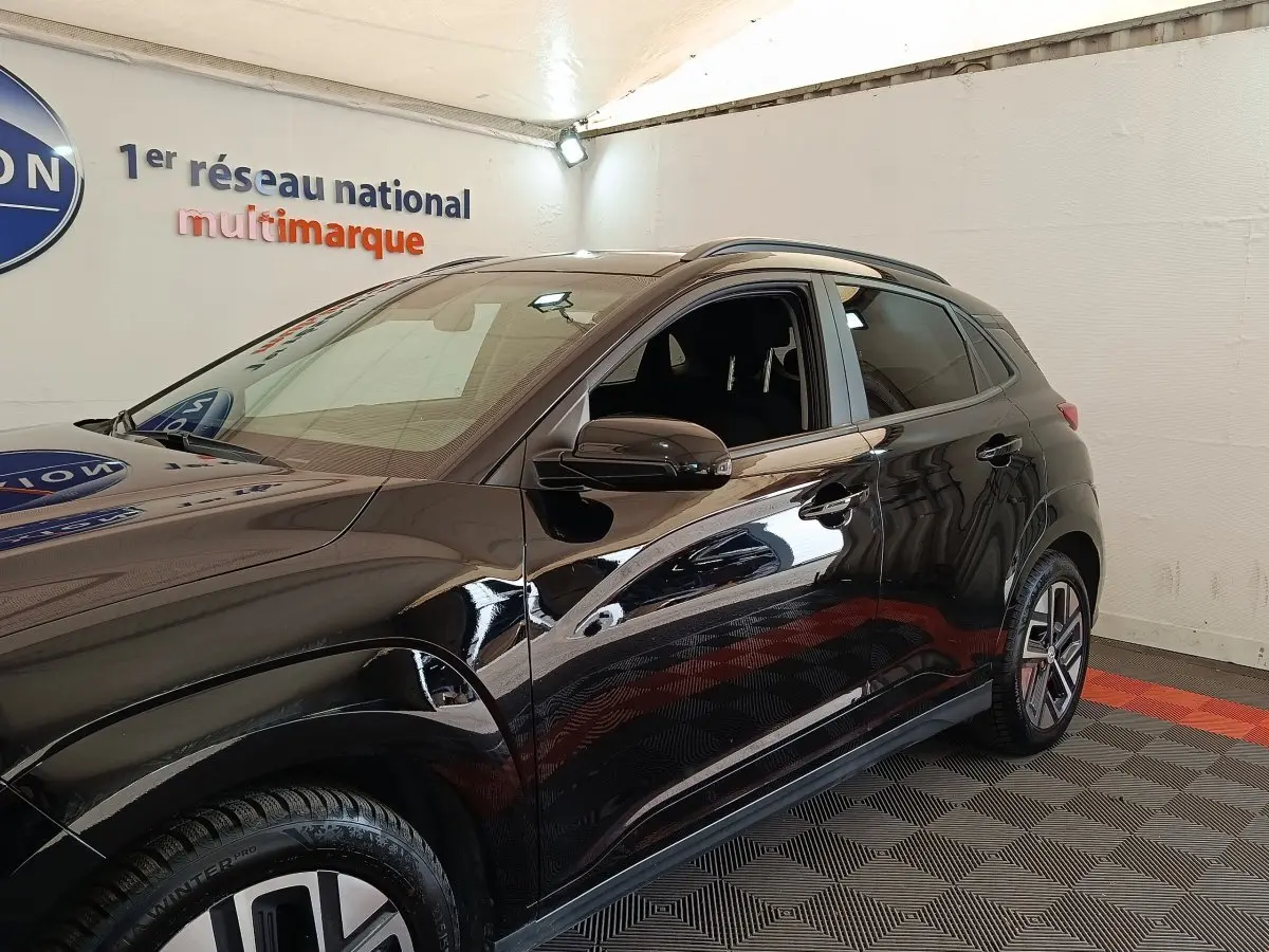 Vue côté gauche du Hyundai Kona électrique noir 2023 dans un showroom avec sol à motifs gris et rouge.