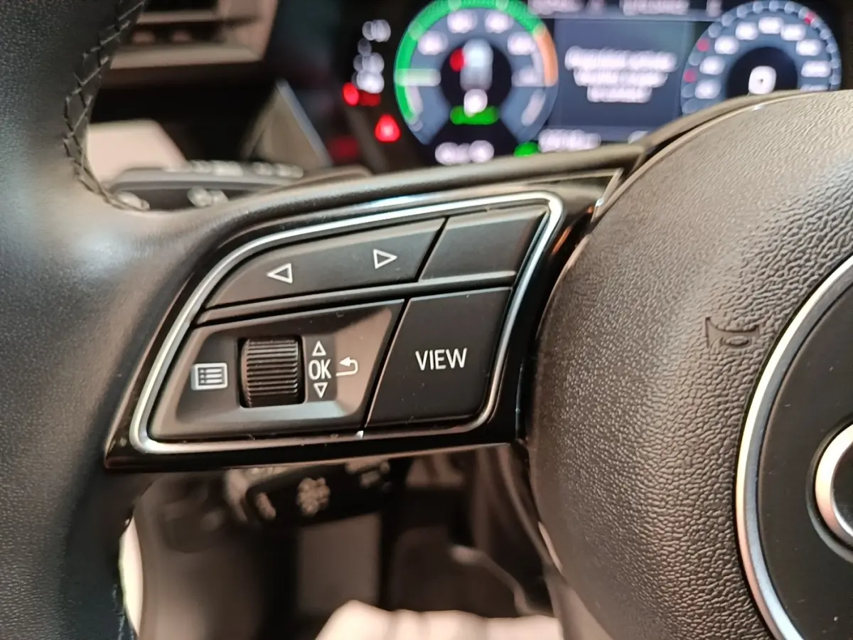 Gros plan sur les commandes gauche du volant cuir noir d’un Audi A3 hybride, avec tableau de bord numérique en arrière-plan flou.